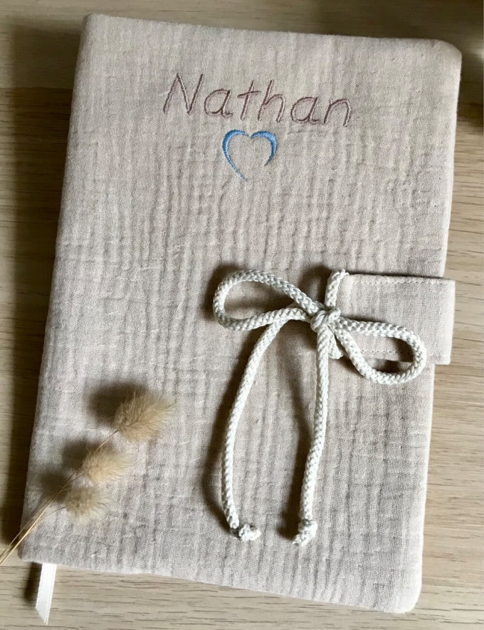 Protège carnet de santé bébé en double gaze de coton lin brodé avec le prénom Nathan et un motif coeur et un cordon.